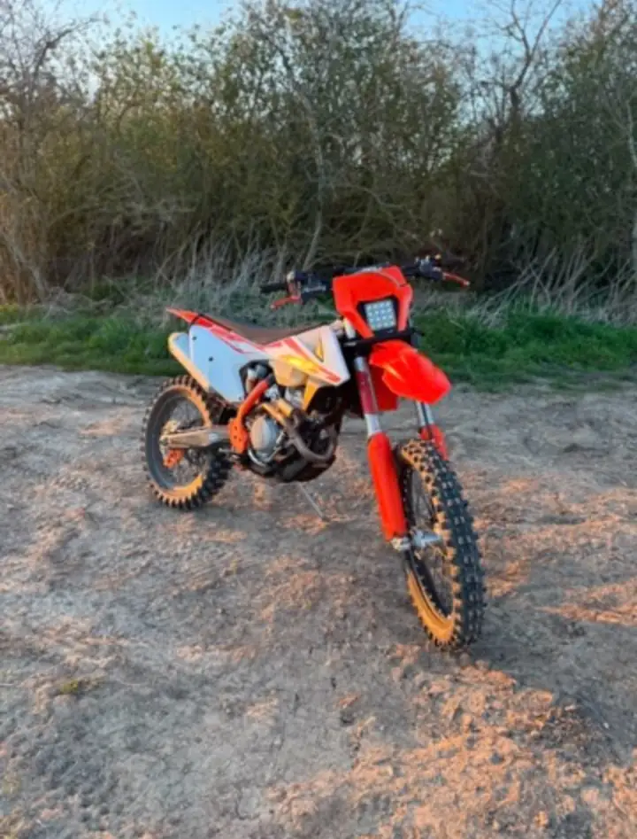 Enduro bike with a square LED headlight and orange forks standing on a sandy track at dusk