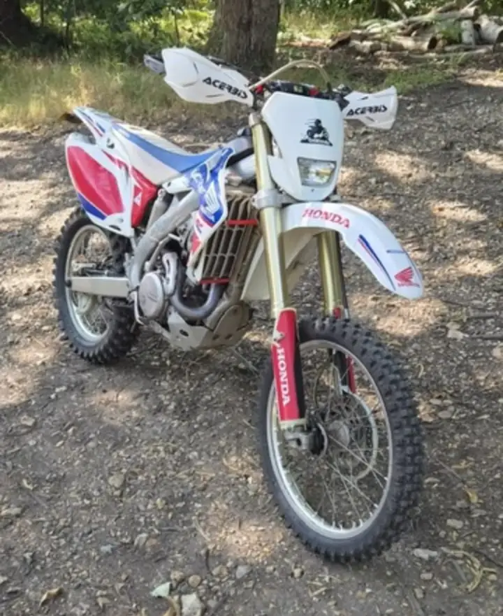 White Honda CRF with red and blue graphics and Acerbis hand guards, parked on a woodland track